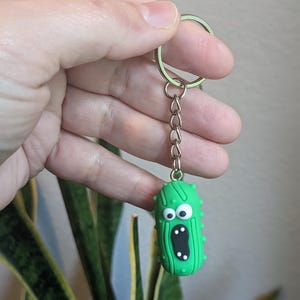 Op de afbeelding: Een groene komkommer sleutelhanger met een verraste uitdrukking en witte ogen. De komkommer heeft een zilveren ketting die aan een zilveren sleutelring is bevestigd.