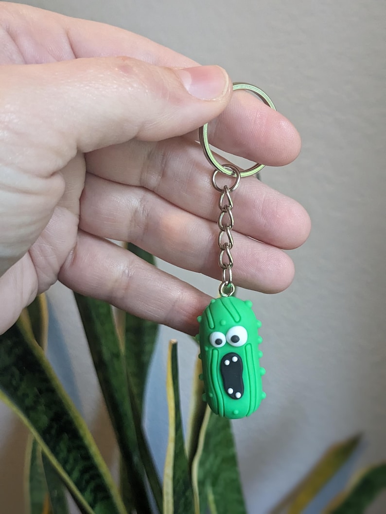 Op de afbeelding: Een groene komkommer sleutelhanger met een verraste uitdrukking en witte ogen. De komkommer heeft een zilveren ketting die aan een zilveren sleutelring is bevestigd.