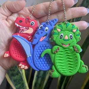 May include: Three colorful dinosaur keychains: a red T-Rex, a blue Pterodactyl, and a green Triceratops. Each keychain is made of a soft, rubbery material and has a silver chain. The dinosaurs have detailed features and are smiling.