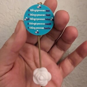 Op de afbeelding: Een blauwe en witte ronde hanger met de tekst "5 dingen die je kunt zien, 4 dingen die je kunt voelen, 3 dingen die je kunt horen, 2 dingen die je kunt ruiken, 1 ding dat je kunt proeven" hangt aan een witte roos aan een bruin touwtje.