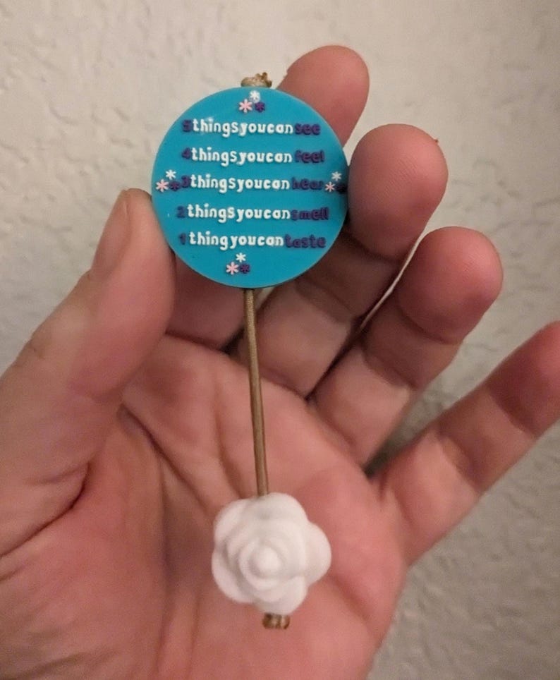 Op de afbeelding: Een blauwe en witte ronde hanger met de tekst "5 dingen die je kunt zien, 4 dingen die je kunt voelen, 3 dingen die je kunt horen, 2 dingen die je kunt ruiken, 1 ding dat je kunt proeven" hangt aan een witte roos aan een bruin touwtje.