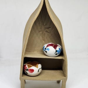 May include: A small, beige ceramic shelf with two shelves. The shelf is shaped like a boat and has a star carved into the top. Two small, white ceramic bowls with painted floral designs are on the shelves.