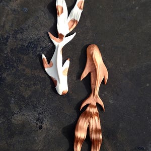 Op de afbeelding: Twee decoratieve koi-vissculpturen. Eén is wit met bruine vlekken en de andere is koperkleurig. De sculpturen zijn gestileerd met vloeiende lijnen en staan tegen een donkere achtergrond.