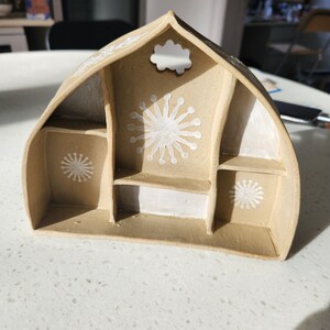 May include: A beige ceramic house-shaped trinket dish with white painted details. The dish has multiple compartments and a small cloud-shaped cutout on the top.