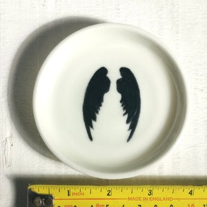 以下が含まれることがあります： 内側に黒い天使の羽根が描かれた、小さな白いセラミック製の皿。皿の直径は約10cmです。