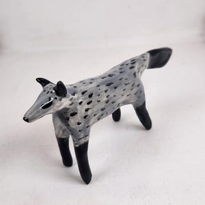 May include: A ceramic fox figurine with a grey body speckled with black spots. The fox has black ears, legs, and tail. The figurine is standing on a white surface.