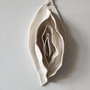 Puede incluir: Escultura de cerámica de una vulva estilizada, con formas en capas en forma de pétalos en tonos blancos y beige. La obra de arte está montada en una pared blanca texturizada, destacando los delicados detalles y las formas orgánicas.