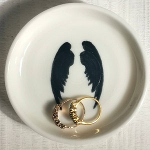 以下が含まれることがあります： 黒い天使の羽のシルエットが描かれた白いセラミック製のリングディッシュ。2つの金のリングがディッシュの中に置かれています。