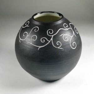 Puede incluir: Un jarrón de cerámica negro con forma redonda y boca ancha. El jarrón presenta un diseño de vid blanca y ondulada alrededor del centro. El interior del jarrón es de color crema claro.