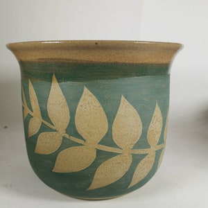 May include: A green ceramic planter with a brown rim and a leaf design. The leaves are a lighter brown color and are arranged in a row around the planter.