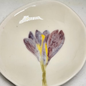 Op de afbeelding: Een klein, crèmekleurig keramiek schaaltje met een handgeschilderd krokusbloem ontwerp. De bloem is afgebeeld in paarse en gele tinten, met een groene stengel. Het schaaltje heeft een onregelmatige, organische vorm.