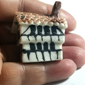 Puede incluir: Una pequeña casa de cerámica blanca con un techo marrón y detalles pintados en negro. La casa tiene una chimenea y dos ventanas.