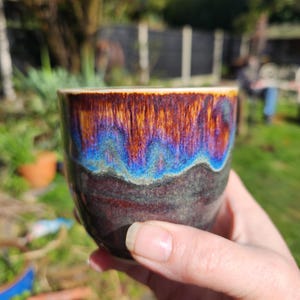 Könnte beinhalten: Eine Keramiktasse mit einer dunkelbraunen, schwarzen und blauen Glasur. Die Tasse hat ein einzigartiges, handgemaltes Design mit einem wellenförmigen Muster.