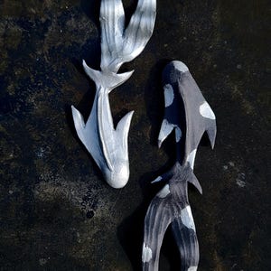 Puede incluir: Dos esculturas decorativas de peces. Una es plateada, la otra es negra con manchas blancas. Ambas tienen un diseño estilizado y abstracto con líneas fluidas y aletas puntiagudas. Las esculturas están colocadas sobre una superficie oscura y texturizada.