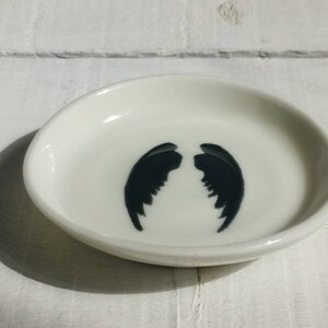 以下が含まれることがあります： 中央に黒い天使の羽のシルエットが描かれた、小さな白いセラミック製の皿です。