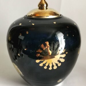 May include: A dark blue ceramic jar with a gold lid and gold accents. The jar has a round shape and is decorated with gold stars and a large gold sunburst design.
