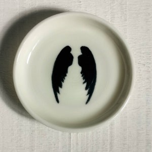 以下が含まれることがあります： 中央に黒い天使の羽のシルエットが描かれた白いセラミック製の皿