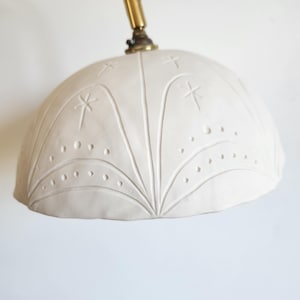 Może przedstawiać: Biały ceramiczny klosz lampy z dekoracyjnym wzorem gwiazd i kropek. Klosz jest przymocowany do złotego metalowego ramienia.