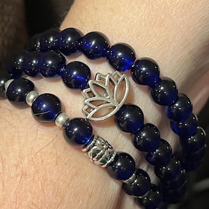 May include: Three dark blue beaded bracelets with silver accents and a silver lotus flower charm. The bracelets are made of round, glossy beads. The lotus charm is in the center of the top bracelet.