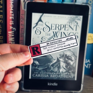May include: A Kindle e-reader displaying the cover of the book "The Serpent & The Wings" by Carissa Broadbent. A red sticker with the letter "R" and the text "RESTRICTED THIS KINDLE CONTAINS SPICY CONTENT (so deliciously its good)" is placed on the screen.