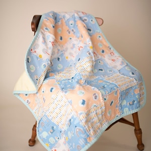 May include: A patchwork baby quilt with a blue, peach, and white color scheme. The quilt features a variety of animal and nature-themed prints. The quilt is folded and draped over a wooden chair.