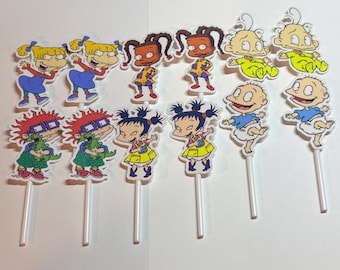 Peut inclure: Une collection de décorations de cupcakes colorées représentant des personnages de dessins animés. Les décorations incluent des personnages aux cheveux blonds, bruns et noirs, portant diverses tenues. Chaque décoration est fixée à un bâtonnet en plastique blanc.