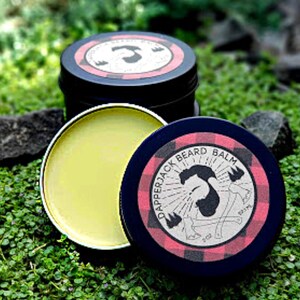 May include: A round tin of beard balm with a red and black plaid design. The label reads "Dapperjack Beard Balm" and features a cartoon illustration of a bearded man with a lumberjack hat and an axe.