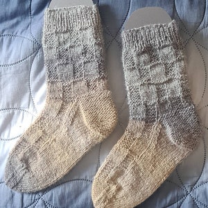 May include: Two pairs of hand-knitted socks in a gradient of grey and beige. The socks have a chequered pattern.