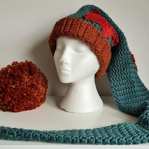 Può includere: Un cappello all'uncinetto blu-verde e ruggine con una lunga coda. Il cappello ha un pon pon all'estremità della coda.