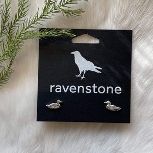 May include: A pair of silver duck earrings on a black card with the word "ravenstone" printed on it.