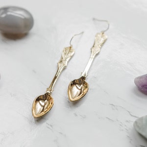 May include: A pair of gold spoon earrings with heart-shaped bowls. The earrings are hanging against a white background with various colored stones.