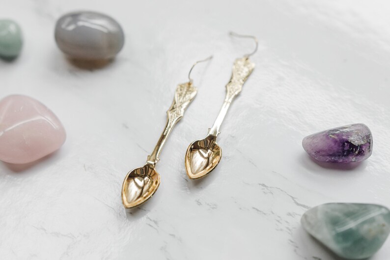 May include: A pair of gold spoon earrings with heart-shaped bowls. The earrings are hanging against a white background with various colored stones.
