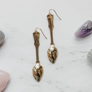 May include: A pair of gold spoon earrings with a long, slender design. The earrings are displayed on a white marble surface with several colorful stones.