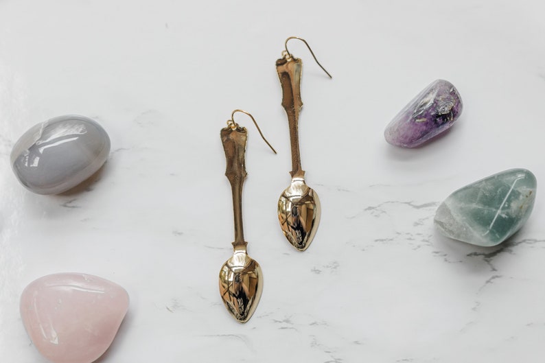 May include: A pair of gold spoon earrings with a long, slender design. The earrings are displayed on a white marble surface with several colorful stones.