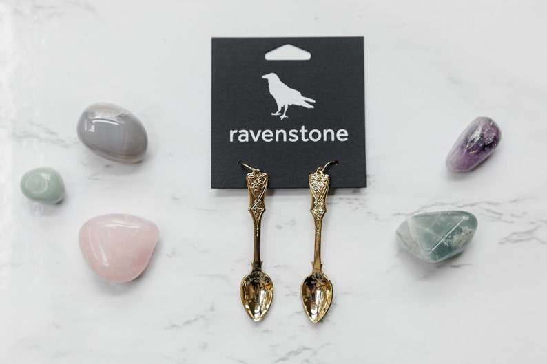 May include: Gold spoon earrings with a decorative design. The earrings are hanging from a black card with the word "ravenstone" printed on it.