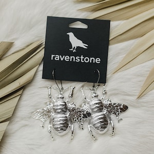 May include: Silver bee earrings with a textured finish. The earrings are on a black card with the text "ravenstone" and a raven logo.