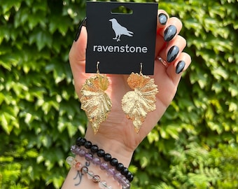 May include: Gold leaf earrings in the shape of a leaf. The earrings are on a black card with the text "ravenstone".