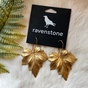 May include: Gold leaf earrings in the shape of maple leaves. The earrings are on a black card with the word "ravenstone" printed on it.