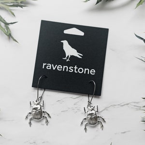 May include: Silver beetle earrings with a black card featuring a white raven and the text "ravenstone".