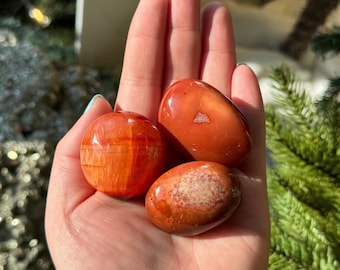 Puede incluir: Tres piedras pulidas, lisas y redondeadas en tonos naranja vibrante y marrón rojizo intenso. Una piedra muestra bandas horizontales distintivas, mientras que las otras presentan superficies sólidas y brillantes con variaciones de color.