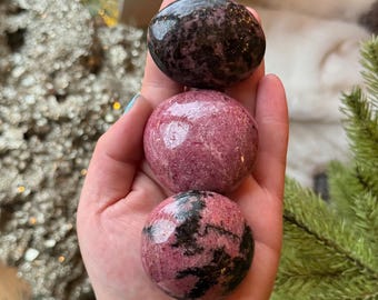 Puede incluir: Tres piedras pulidas y redondeadas apiladas verticalmente. La piedra superior es de color marrón oscuro con motas negras. La piedra central es de un rosa vibrante y sólido con un acabado liso y brillante. La inferior es rosa con vetas negras.