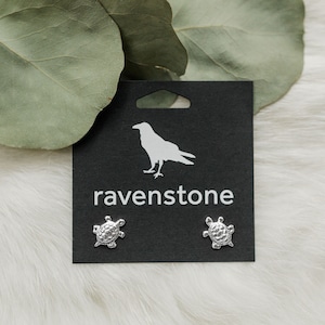 May include: A pair of silver turtle stud earrings on a black card with the ravenstone logo. The card has a hole at the top for hanging.
