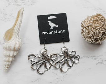 May include: Silver-colored octopus earrings with a dangling hook design. The earrings are displayed on a black card with the word "ravenstone" and a raven logo. A white seashell and a desert rose are also in the image.