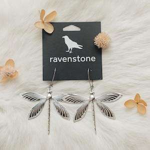 May include: Silver dragonfly earrings with intricate details, hanging from a black card with the text "ravenstone" and a silhouette of a raven.
