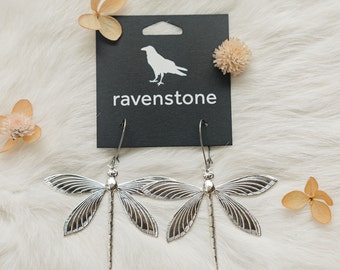 Puede incluir: Pendientes de libélula de plata con detalles intrincados, colgando de una tarjeta negra con el texto "ravenstone" y una silueta de un cuervo.
