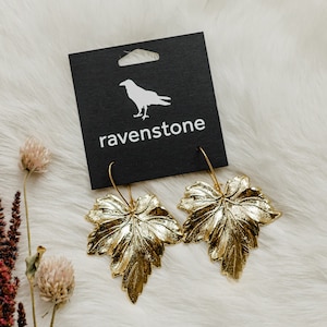 May include: Gold leaf earrings shaped like maple leaves. The earrings are on a black card with the word "ravenstone" printed on it.