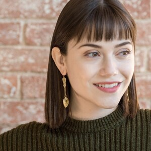 May include: A woman with short brown hair wearing a green sweater and gold spoon earrings. She is smiling and looking to the right.