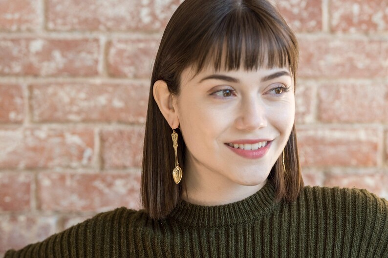 May include: A woman with short brown hair wearing a green sweater and gold spoon earrings. She is smiling and looking to the right.