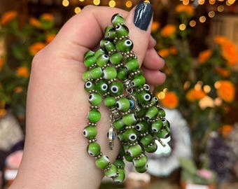 May include: A strand of green beaded jewelry with white and black eye-shaped beads. The beads are strung together with silver-colored metal. A silver charm with a blue eye is attached to the strand. The background is blurred with orange and green foliage.