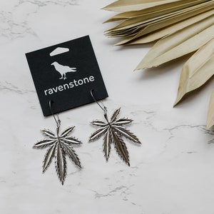 May include: Silver marijuana leaf earrings on a white background with a black card that says "ravenstone" and a small bird graphic.